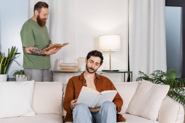 Gay man reading book near partner in living room at home  - 写真・画像