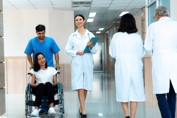 Doktor ve erkek hemşire, steril hastane koridorunda tekerlekli sandalyeli bir kadın hastayı naklediyor. Engelli hastalar için hastane konseptinde sağlık ve hemşirelik hizmeti. - Fotoğraf, Görsel