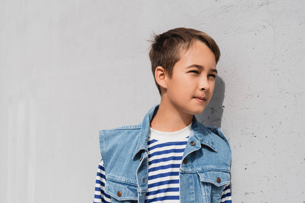 well dressed preteen boy in denim vest and striped long sleeve shirt standing near mall with grey wall - Фото, изображение