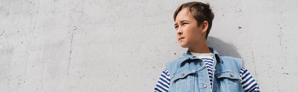 low angle view of stylish boy in denim vest and striped long sleeve shirt looking away near mall with grey wall, banner  - Foto, immagini