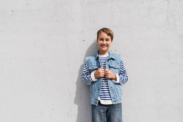 garçon heureux en gilet en denim et chemise à manches longues rayée debout près du centre commercial avec mur gris - Photo, image