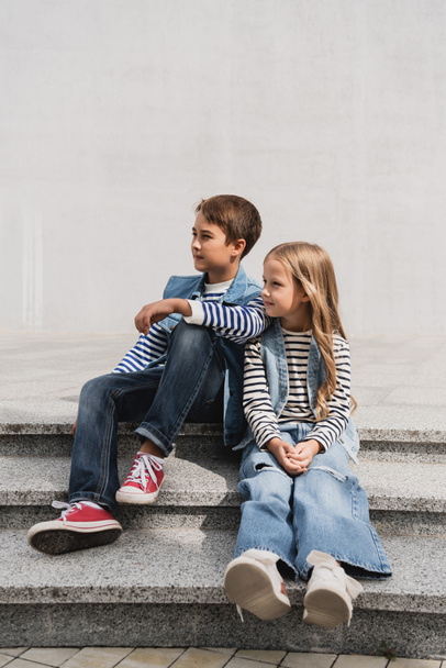 full length of well dressed children in denim outfits sitting on stairs near building  - Foto, Imagen