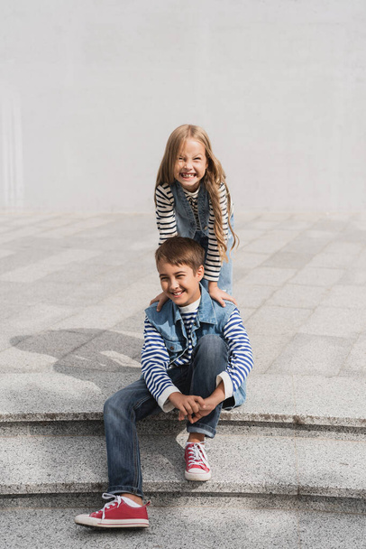 cheerful girl in stylish clothes leaning on shoulders of happy preteen boy sitting on stairs  - Foto, Bild