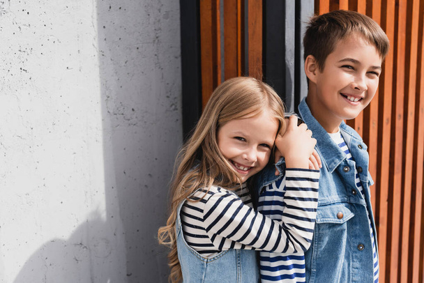 portrait of cheerful kids in striped long sleeve shirts and denim vests looking at camera near building  - Фото, изображение