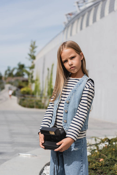 preteen girl in denim vest and striped long sleeve shirt holding vintage camera near mall building  - Foto, Imagen