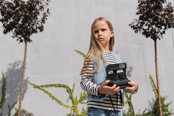 girl in blue denim vest and striped long sleeve shirt holding vintage camera near mall building  - Foto, afbeelding
