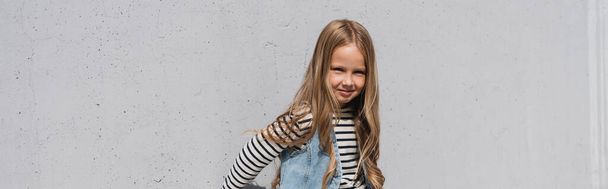 cheerful girl in denim vest striped long sleeve shirt standing near grey wall, banner   - Φωτογραφία, εικόνα