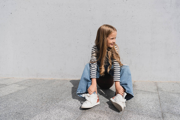 full length of smiling girl in denim vest and blue jeans sitting on basketball near wall  - Photo, Image