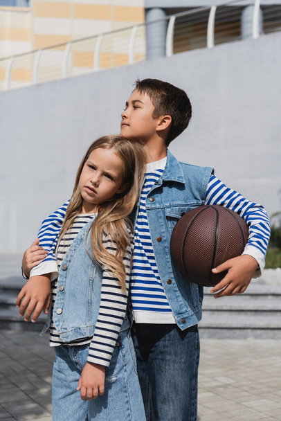хлопчик в джинсовому жилеті тримає баскетбол і обіймає стильну дівчину, стоячи біля торгового центру
  - Фото, зображення