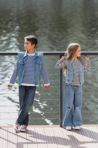 full length of well dressed kids in denim vests and jeans posing next to metallic fence on river embankment - Foto, afbeelding