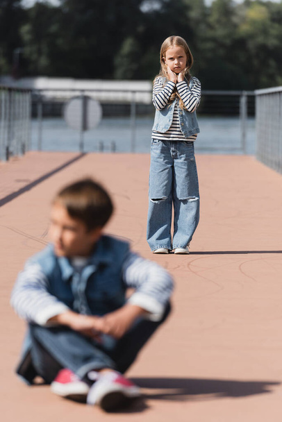 full length of preteen girl in denim outfit standing on river embankment near blurred boy on foreground  - Zdjęcie, obraz