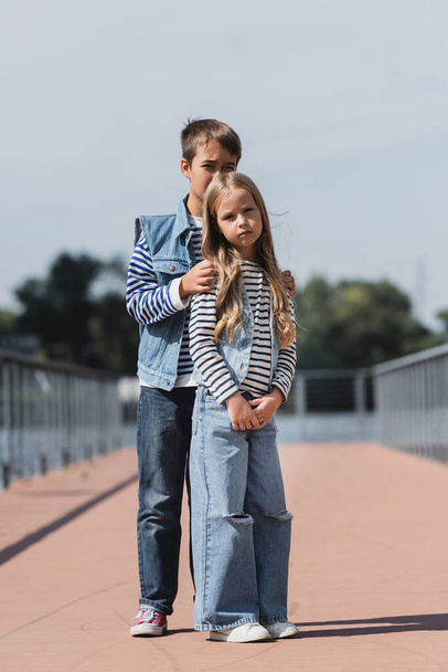 full length of preteen boy and girl in denim clothes standing on riverside embankment  - Foto, Bild