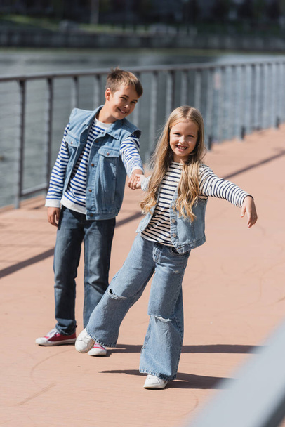 longitud completa de niños felices en ropa de mezclilla tomados de la mano mientras se divierten en el terraplén del río  - Foto, Imagen