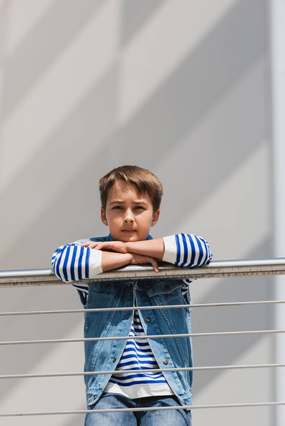 stylish boy in denim vest and long sleeve shirt standing near metallic fence on embankment  - 写真・画像