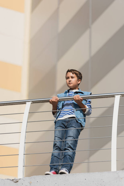 vue à angle bas du garçon élégant en gilet en denim et jeans debout près d'une clôture métallique  - Photo, image
