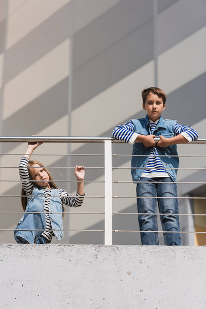 low angle view of children in stylish denim vests and striped long sleeve shirts standing near metallic fence  - Foto, Imagen