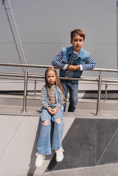 stylish kids in casual denim attire posing near metallic handrails next to building  - Foto, afbeelding