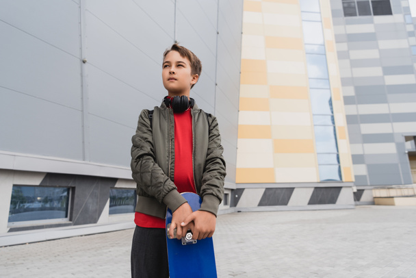 preteen boy in bomber jacket and wireless headphones holding penny board while looking away near mall  - Fotoğraf, Görsel