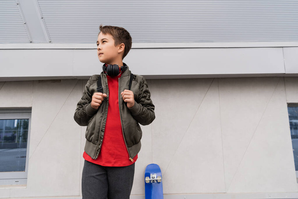 preteen boy in bomber jacket and wireless headphones standing with backpack near penny board  - Zdjęcie, obraz