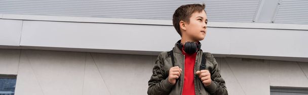 preteen boy in bomber jacket and wireless headphones holding backpack while standing near mall, banner  - Photo, image
