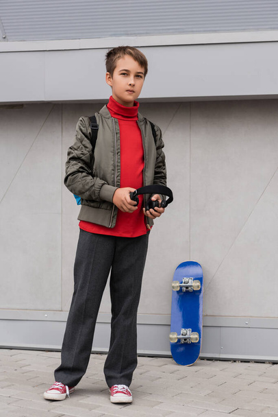 preteen boy in bomber jacket holding wireless headphones while standing near penny board next to mall  - Photo, Image