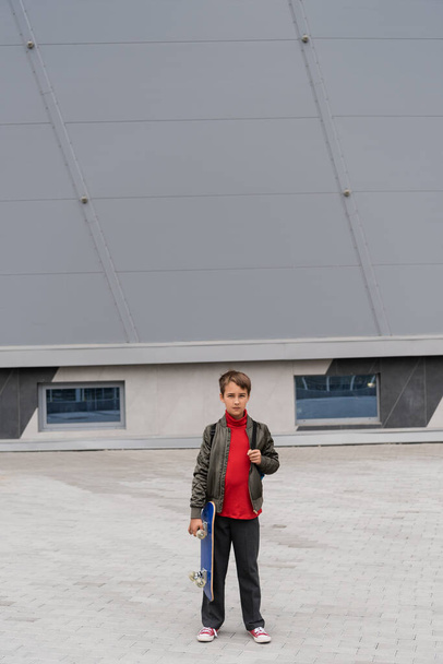 full length of preteen boy in stylish bomber jacket holding penny board near mall building  - Photo, Image