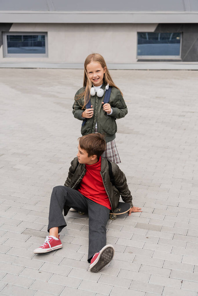 full length of preteen boy in trendy bomber jacket sitting on penny board near cheerful girl  - Photo, image