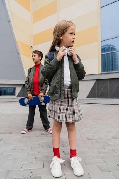 full length of preteen girl in wireless headphones standing with stylish boy holding penny board on blurred background  - Zdjęcie, obraz