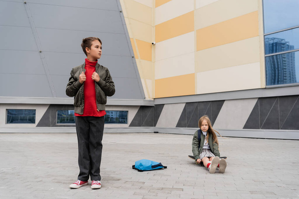 preteen girl in skirt sitting on penny board near stylish boy in trendy bomber jacket next to mall  - Foto, Bild