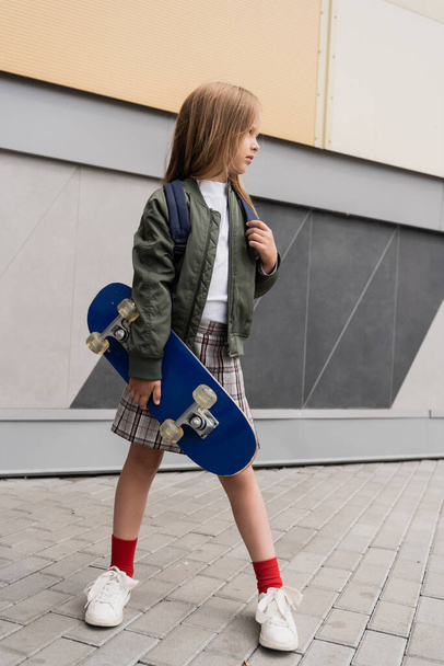 full length of stylish preteen girl in bomber jacket holding penny board standing near mall  - Foto, imagen
