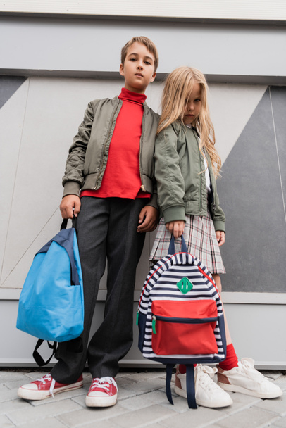 low angle view of stylish kids in bomber jackets holding backpacks while standing near mall  - Φωτογραφία, εικόνα
