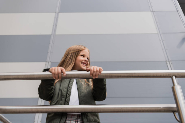 low angle view of cheerful preteen girl in bomber jacket leaning on metallic handrails near mall  - 写真・画像
