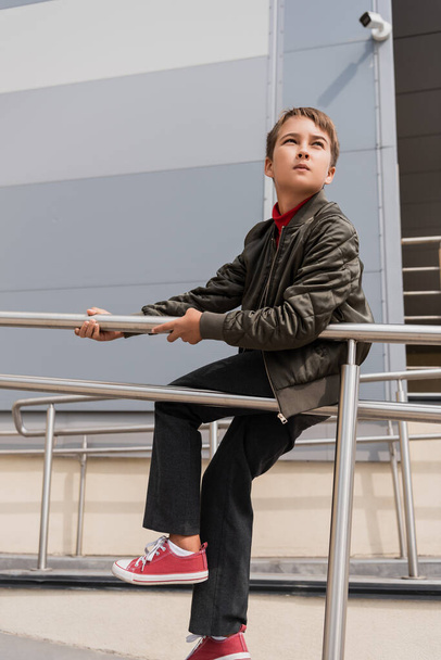 preteen boy in stylish bomber jacket sitting on metallic handrails near mall  - Valokuva, kuva