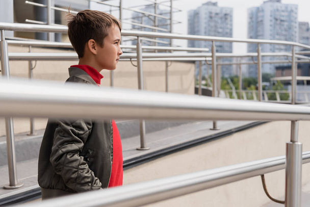 well dressed preteen boy in stylish bomber jacket standing near metallic handrails on blurred foreground  - Foto, Bild
