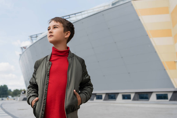well dressed preteen boy in stylish bomber jacket and red turtleneck posing with hands in pockets near mall  - Foto, imagen