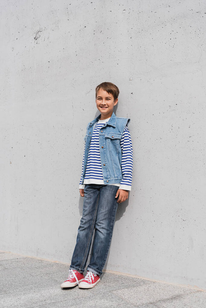 full length of happy boy in striped long sleeve shirt and denim vest leaning on wall outdoors  - Fotó, kép