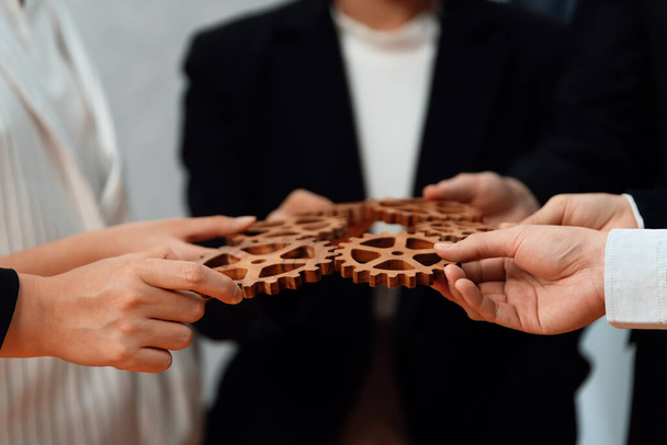 Closeup hand holding wooden gear by businesspeople wearing suit for harmony synergy in office workplace concept. Group of people hand making chain of gears into collective form for unity symbol. - Foto, Imagem