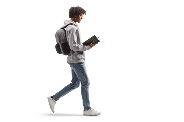 Full length profile shot of a young african american man walking and reading a book isolated on white background - Photo, Image