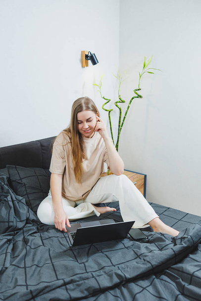 Mujer joven sonriente con ropa casual sonriendo y trabajando a distancia, sentada en la cama con el ordenador portátil durante el trabajo remoto. Trabajo independiente. Trabajo desde casa - Foto, imagen