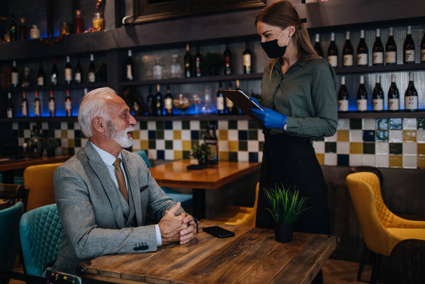 Waitress serves and takes the order from the senior businessman at the restaurant. She wears a protective mask as part of security measures against the Coronavirus pandemic. - Φωτογραφία, εικόνα