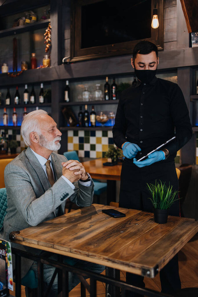 The waiter taking order for a delicious meal to the senior businessman at the restaurant. He wears a protective mask as part of security measures against the Coronavirus pandemic. - Foto, afbeelding