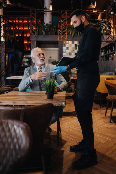 The waiter taking order for a delicious meal to the senior businessman at the restaurant. He wears a protective mask as part of security measures against the Coronavirus pandemic. - Foto, immagini