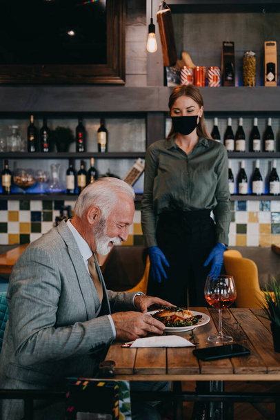Waitress serves and takes the order from the senior businessman at the restaurant. She wears a protective mask as part of security measures against the Coronavirus pandemic. - Photo, Image