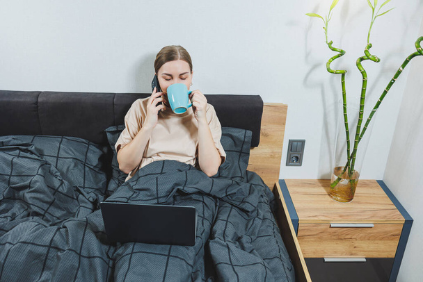 若い美しい女性がコーヒーとベッドの上に座って、自宅でコンピュータを操作し、リモートワーク。女性幸せと笑顔幸せなベッドで自宅. - 写真・画像