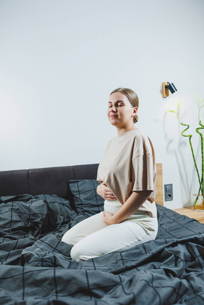 Roztomilá mladá těhotná žena sedící na posteli s šedými prostěradly v neformálním oblečení. Roztomilá těhotná žena doma. - Fotografie, Obrázek