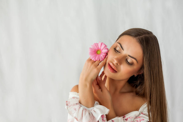 Portrait of young beautiful long-haired brown-eyed woman with pink gerbera on white background. Spring. Holidays. - Foto, afbeelding