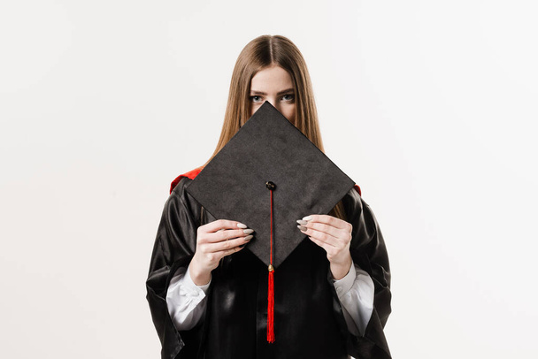 Absolvent dívka magisterský titul v černé maturitní šaty je držení čepice v rukou na bílém pozadí. Atraktivní mladá žena absolvovala vysokou školu - Fotografie, Obrázek