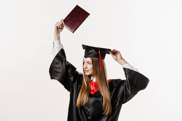 Happy girl student in black graduation gown and cap raises masters degree diploma above head on white background. Graduate girl is graduating college and celebrating academic achievement - Photo, Image