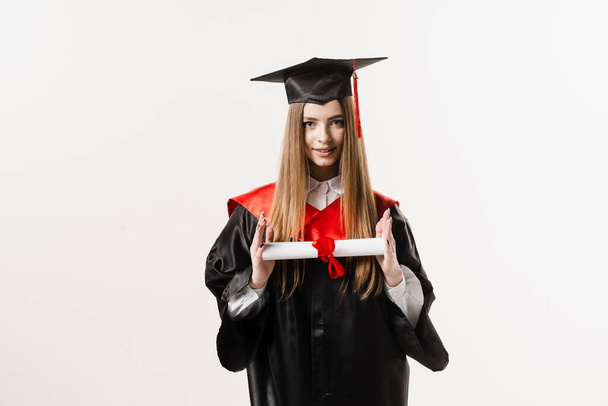Graduate girl with master degree diploma in black graduation gown and cap on white background. Happy young woman successfully graduated from the university with honors - Foto, afbeelding