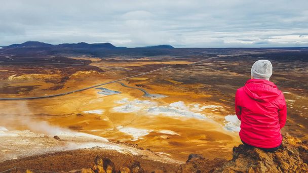 Dziewczyna ubrana w różową kurtkę siedzi na szczycie góry błota, z widokiem na geotermalną plamę znaną z bulgoczących basenów błota i parujących fumaroli emitujących gaz siarkowy. Ekstremalny krajobraz. - Zdjęcie, obraz